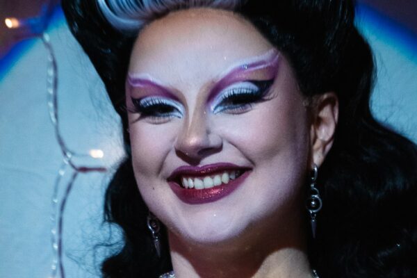 Médée The Bwitch en gros plan, maquillage burlesque graphique rose et blanc, coiffure noire rétro avec mèche blanche, souriante sur scène, photographiée par Miko Michto.