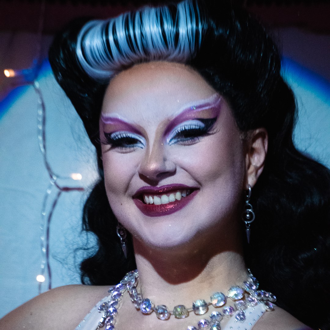 Médée The Bwitch en gros plan, maquillage burlesque graphique rose et blanc, coiffure noire rétro avec mèche blanche, souriante sur scène, photographiée par Miko Michto.