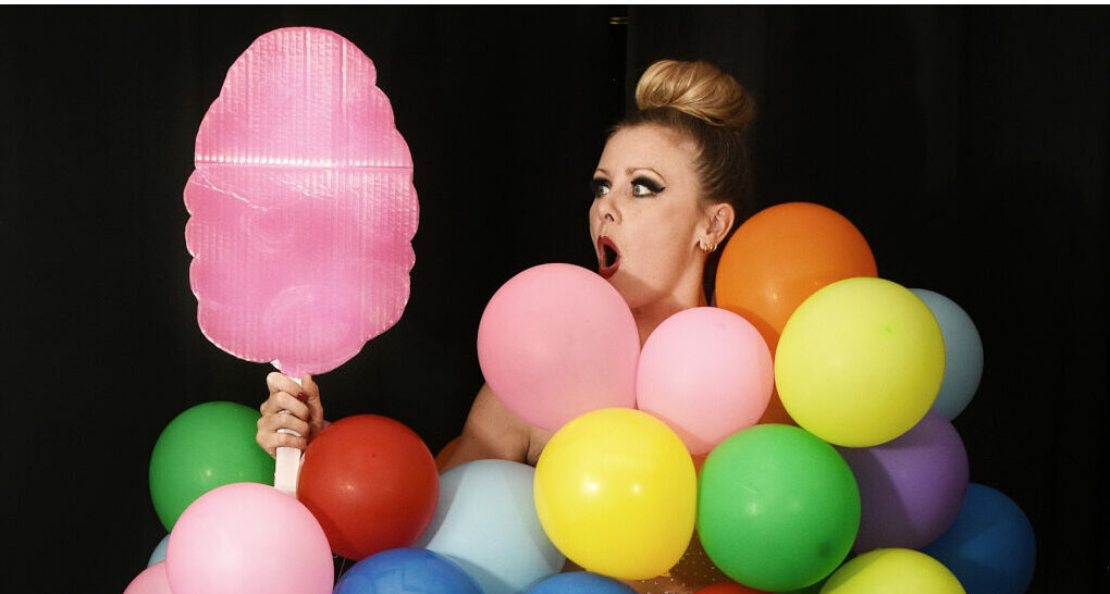 Artiste burlesque Lara Broca recouverte de ballons multicolores, tenant un accessoire rose en forme de barbe à papa devant un fond noir.