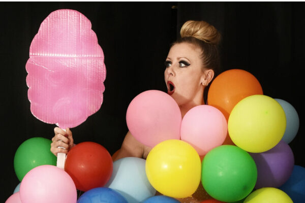 Artiste burlesque Lara Broca recouverte de ballons multicolores, tenant un accessoire rose en forme de barbe à papa devant un fond noir.
