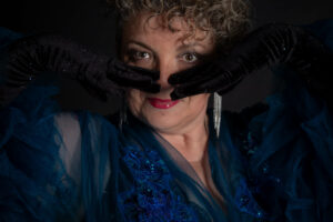 Lady Poppy Scarlett, artiste burlesque, cheveux bouclés blancs, gants noirs couvrant visage, robe bleue brodée. Photographe : Cécile Schwaller.