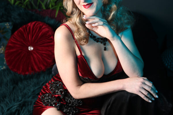 Magali Bertine robe rouge fendu velours plumes tropicales, boucles blondes collier perles. Photographe : Pinup Addict.