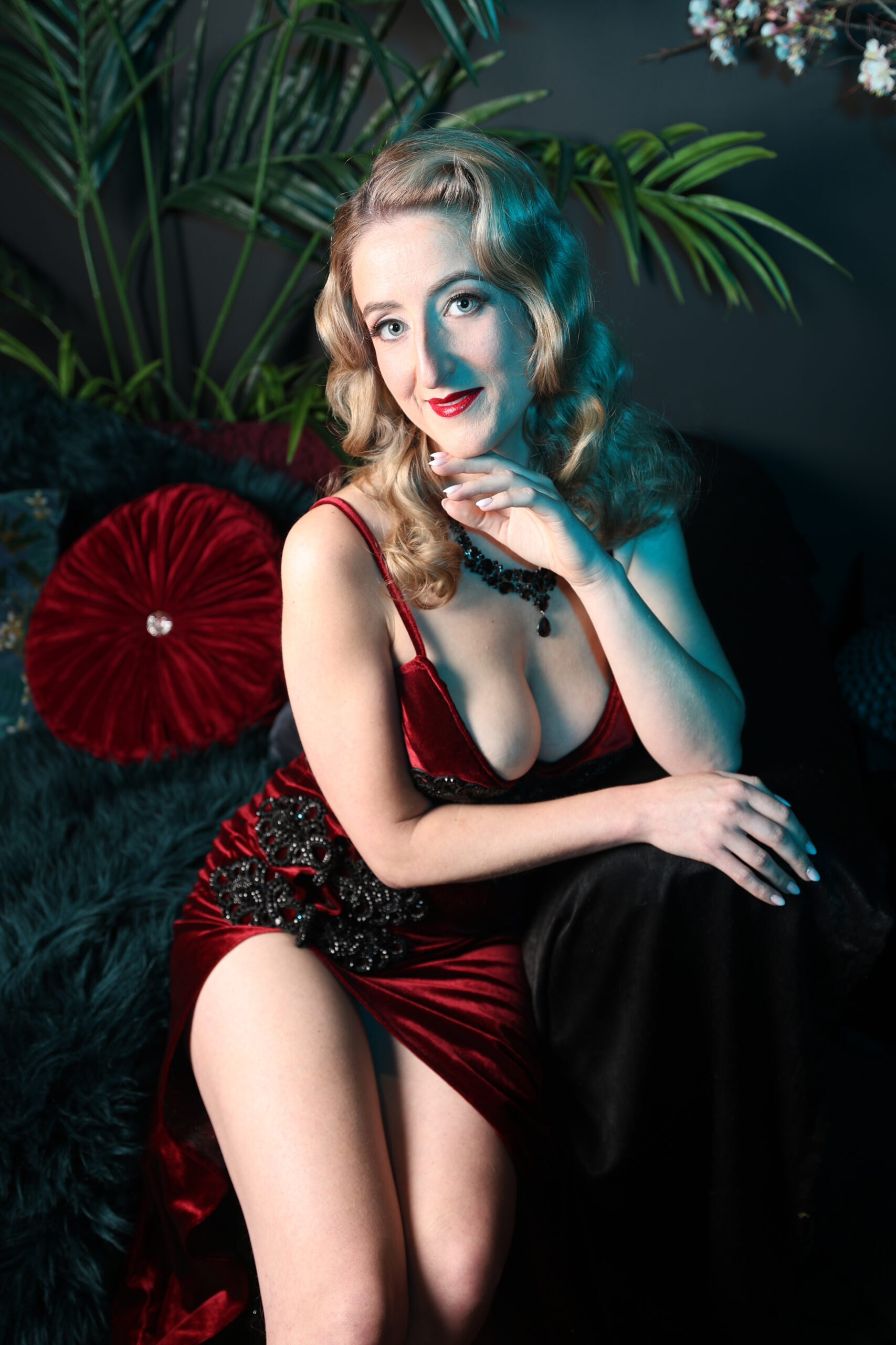Magali Bertine robe rouge fendu velours plumes tropicales, boucles blondes collier perles. Photographe : Pinup Addict.