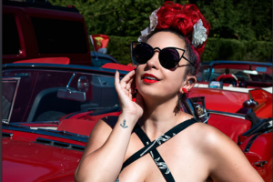 Nidcy Namite, artiste burlesque pin-up, cheveux rouges fleuris lunettes soleil, robe noire imprimé feuilles voiture rouge vintage. Photographe : Fairy Decadence.