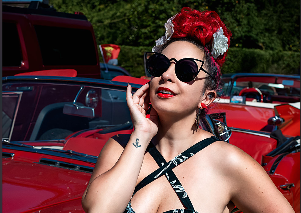 Nidcy Namite, artiste burlesque pin-up, cheveux rouges fleuris lunettes soleil, robe noire imprimé feuilles voiture rouge vintage. Photographe : Fairy Decadence.