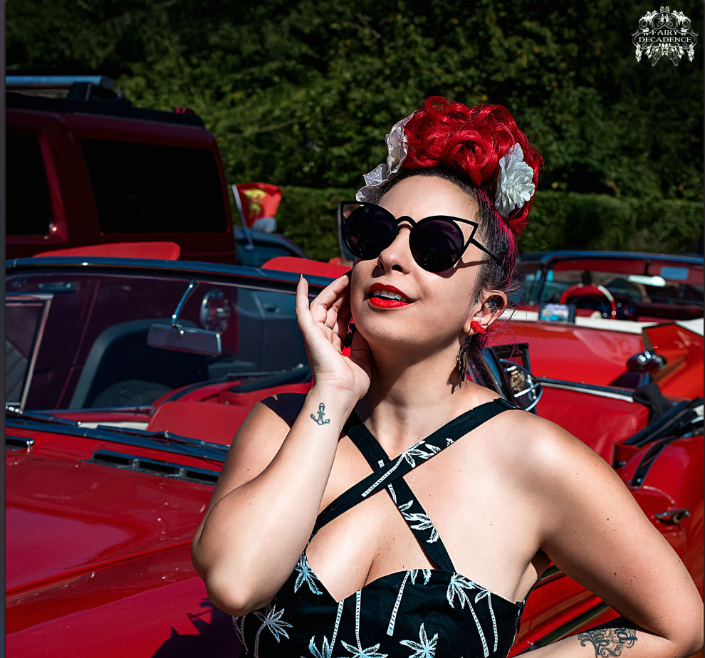 Nidcy Namite, artiste burlesque pin-up, cheveux rouges fleuris lunettes soleil, robe noire imprimé feuilles voiture rouge vintage. Photographe : Fairy Decadence.
