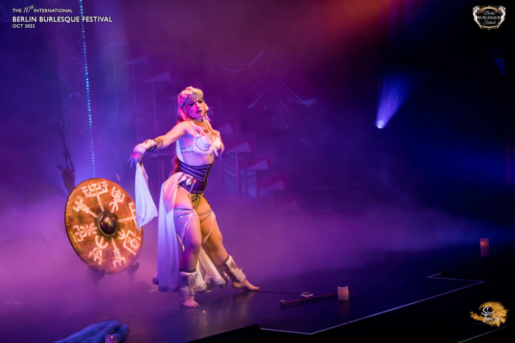 Daâr Djiling sur scène festival burlesque en guerrière viking avec bouclier hache tunique blanche éclairage violet