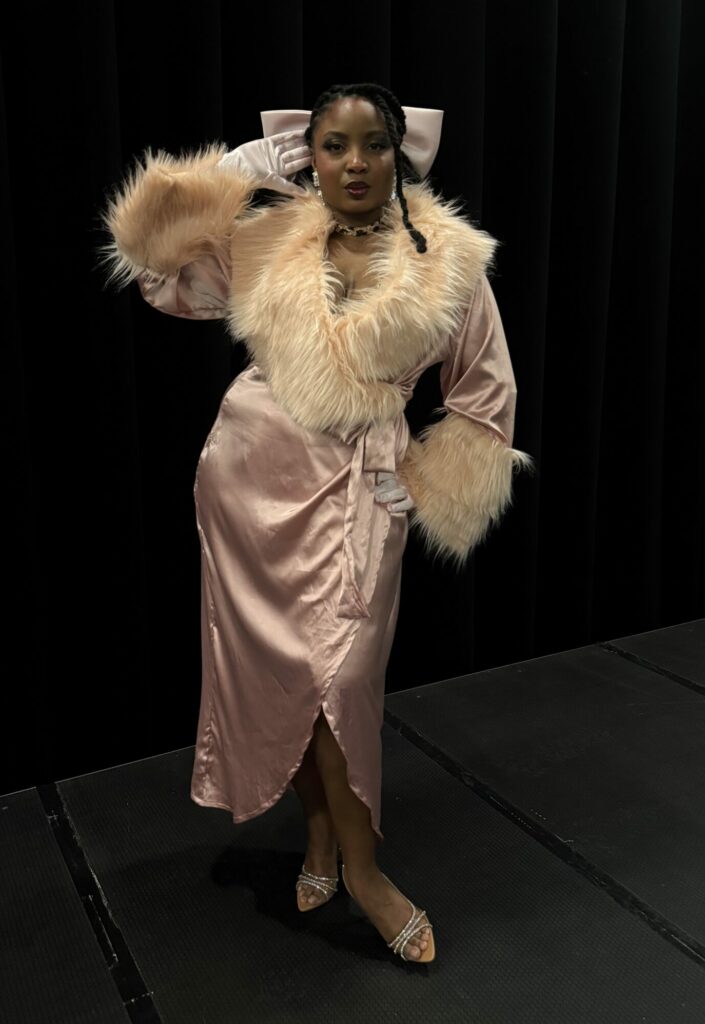 Nubsaphrodite, performer burlesque, en peignoir rose fourré et nœud tête, pose sur scène sombre.