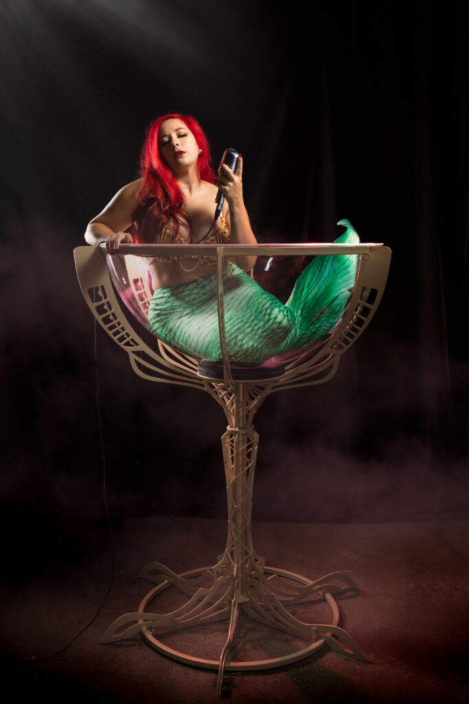 Nidcy Namite en sirène burlesque verre géant, queue écaille verte lingerie or, micro main, fumée lumière rouge. Photographe : Sushi Photo Lab.