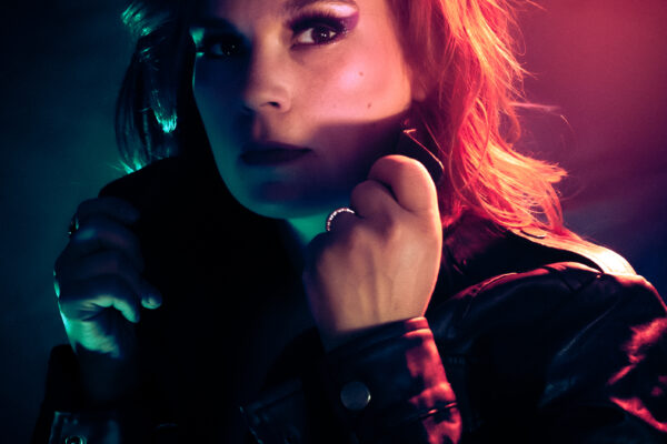 Patti Bear, artiste burlesque rousse, veste cuir noire éclairage néon violet rose, regard intense. Photographe : Jubadi Photography