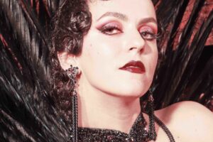 Portrait Velena Rossa style rétro 1920s, coiffure ondulée noire collier chaînes noires lèvres rouges. Crédit photo : Neil Kendall