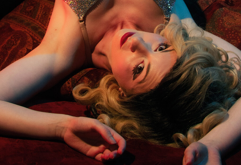 Portrait d'Eden Weiss, artiste burlesque aux cheveux blonds ondulés, en pose glamour sur fond rouge.