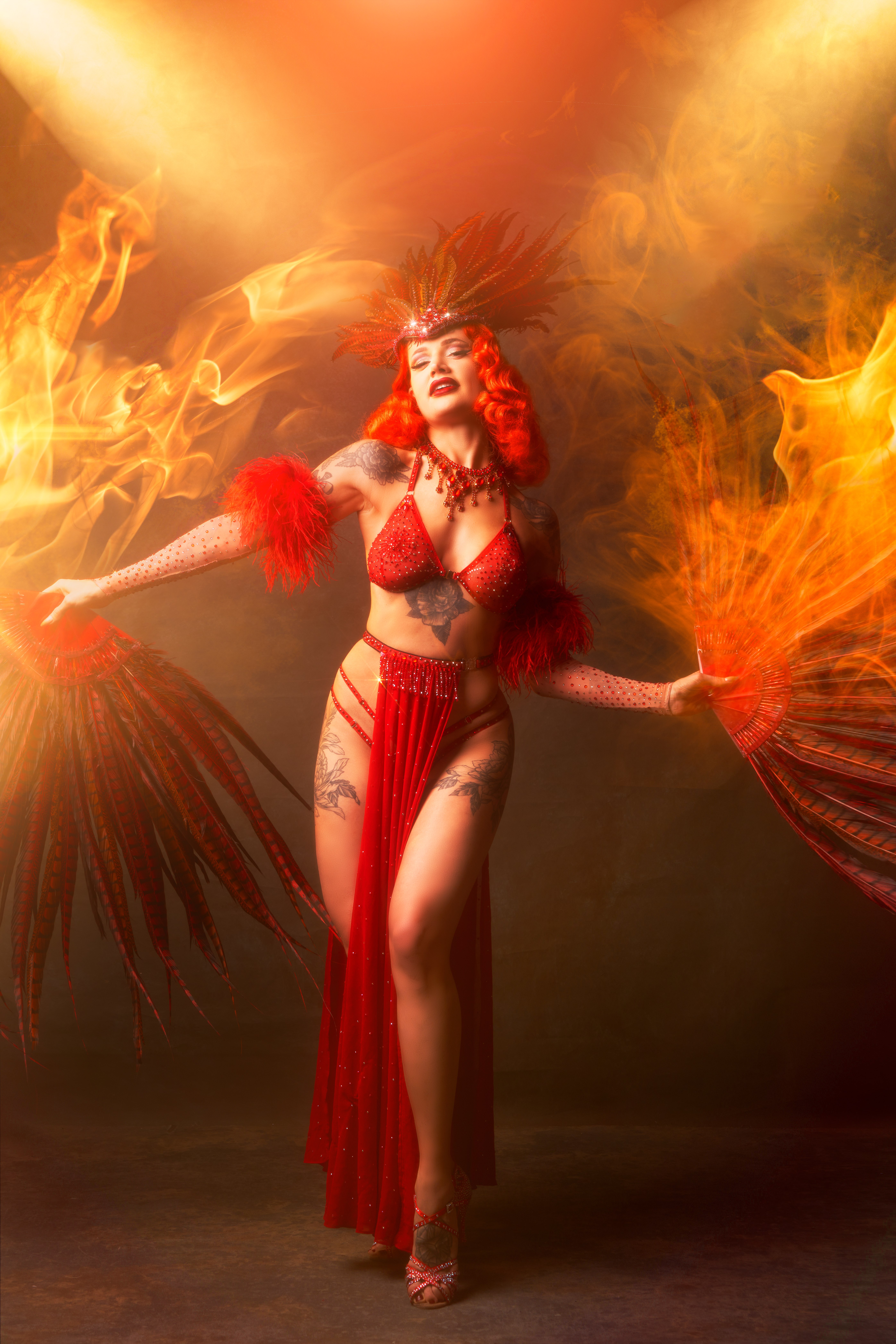 Bettie Rebelle sur scène en costume de scène rouge étincelant avec des éventails en plumes, entourée d'effets de flammes. Photo par Mathys Fine Art.